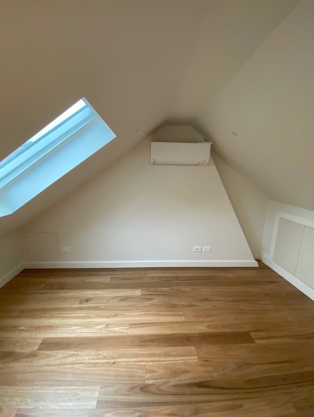 Pomalowane na biało poddasze ze skośnym oknem dachowym, klimatyzatorem i drewnianą podłogą w odcieniach brązu.