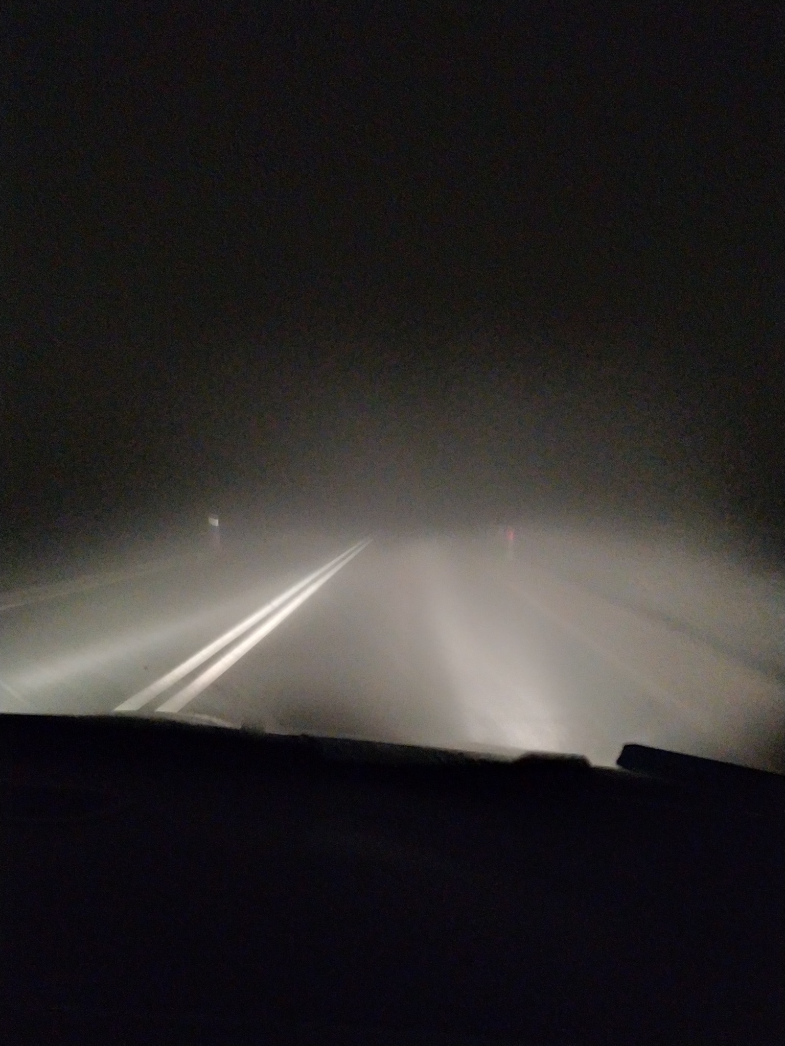 Widok z wnętrza samochodu na drogę w gęstej mgle, oświetloną światłami pojazdu, widoczne podwójne linie na jezdni i słabo widoczny znak drogowy w oddali.