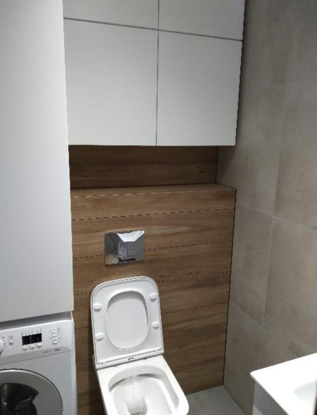 Nowoczesna toaleta podwieszana z deską wolnoopadającą, wkomponowana w zabudowę z drewnopodobnych paneli ściennych, obok pralka i szafki wiszące.