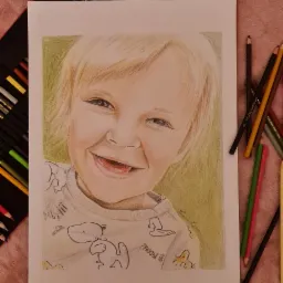 Portret dziecka narysowany kredkami na formacie A3, widoczny na tle z rozłożonym zestawem kredek i kilkoma kredkami leżącymi obok rysunku. Dziecko uśmiechnięte, ubrane w bluzkę z motywem Snoopy.