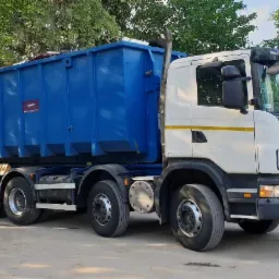 Ciężarówka Scania z niebieskim kontenerem na gruz, zaparkowana na placu, widoczne tablice rejestracyjne.