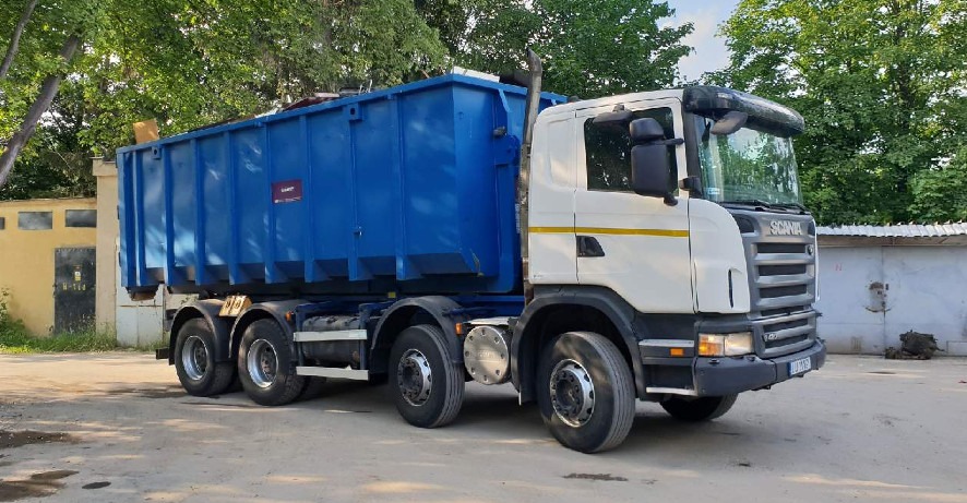Ciężarówka Scania z niebieskim kontenerem na gruz, zaparkowana na placu, widoczne tablice rejestracyjne.