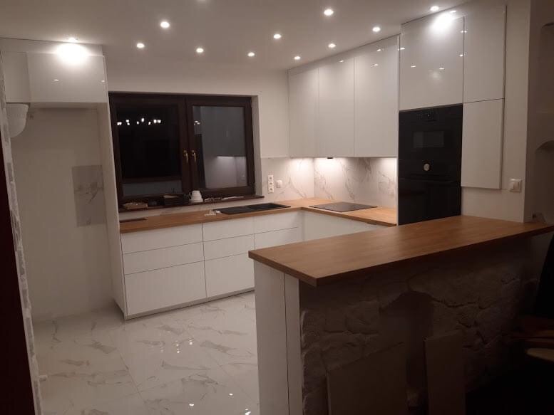 Nowoczesna kuchnia z białymi, lakierowanymi frontami szafek, drewnianymi blatami i oświetleniem punktowym, widziana z perspektywy baru wykończonego dekoracyjną cegłą.