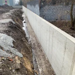 Wykop pod fundament z częściowo wylanym betonem, widoczne ślady po deskowaniu. W tle budynek mieszkalny i szary mur z bloczków betonowych.