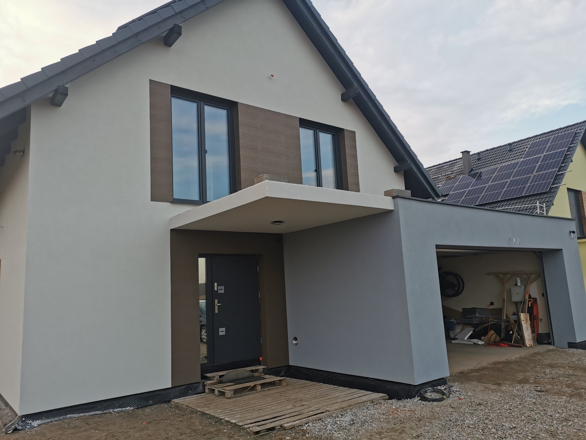 Nowoczesny dom jednorodzinny z szarą elewacją, brązowymi akcentami przy oknach i garażem z otwartą bramą, widoczne przygotowania do zagospodarowania terenu wokół domu.