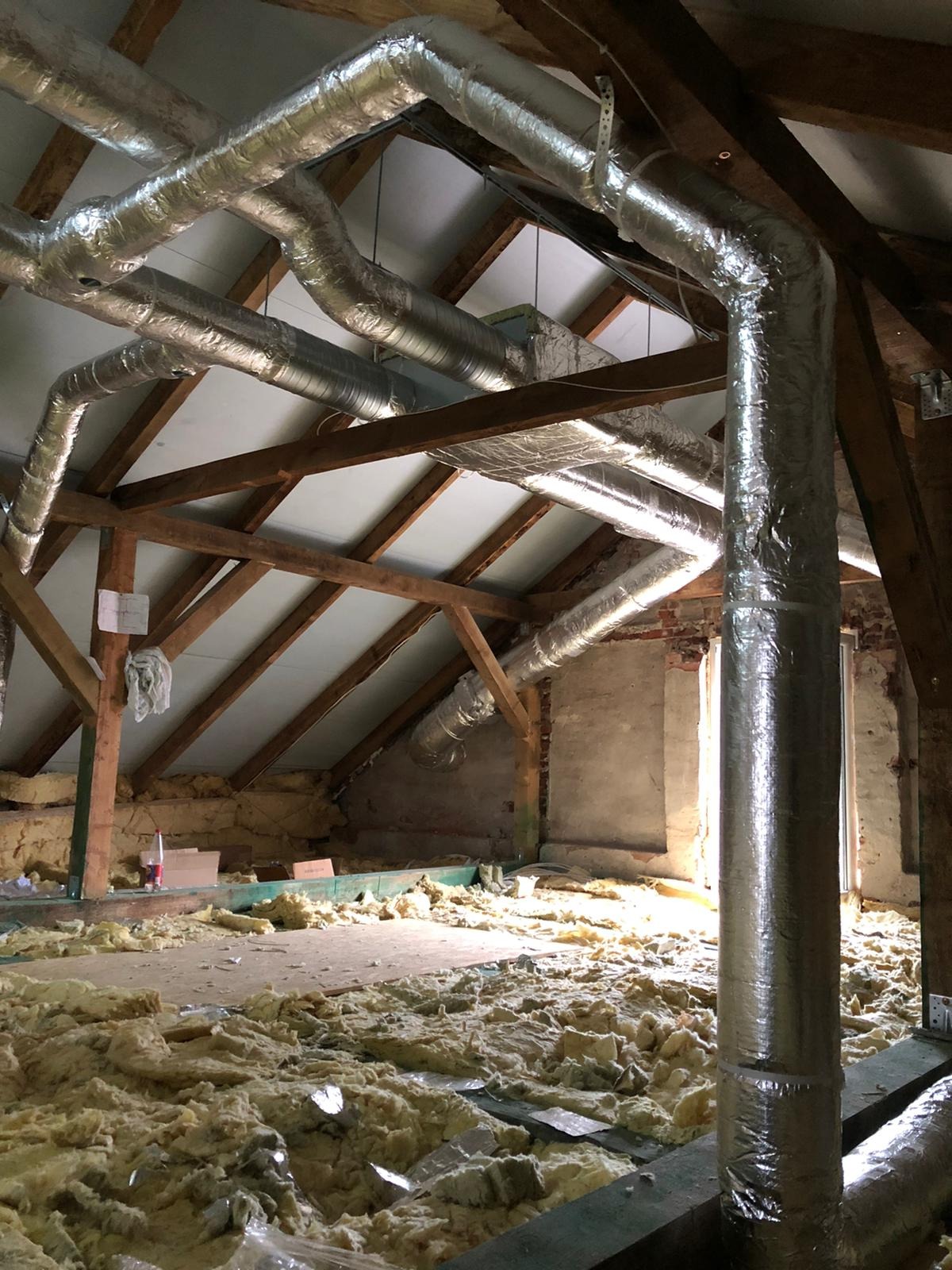 Instalacja przewodów wentylacyjnych z izolacją termiczną w surowym stanie poddasza z widoczną drewnianą konstrukcją dachu i oknem w tle.