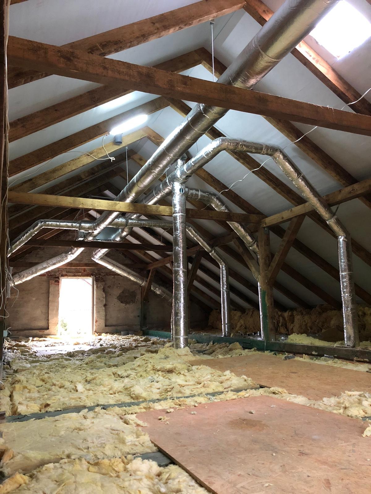 Montaż srebrnych rur wentylacyjnych w przestrzeni poddasza z drewnianą konstrukcją i częściowo ułożoną izolacją z wełny mineralnej.