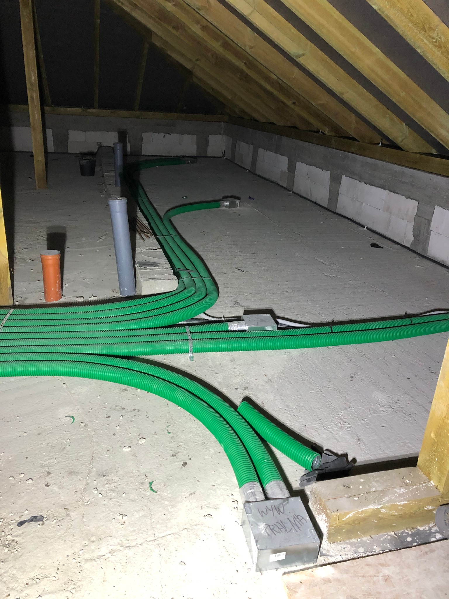 Zielone, elastyczne rury wentylacyjne ułożone na poddaszu, przymocowane metalowymi zaciskami, widoczne skrzynki przyłączeniowe i fragmenty ścian z bloczków betonowych.