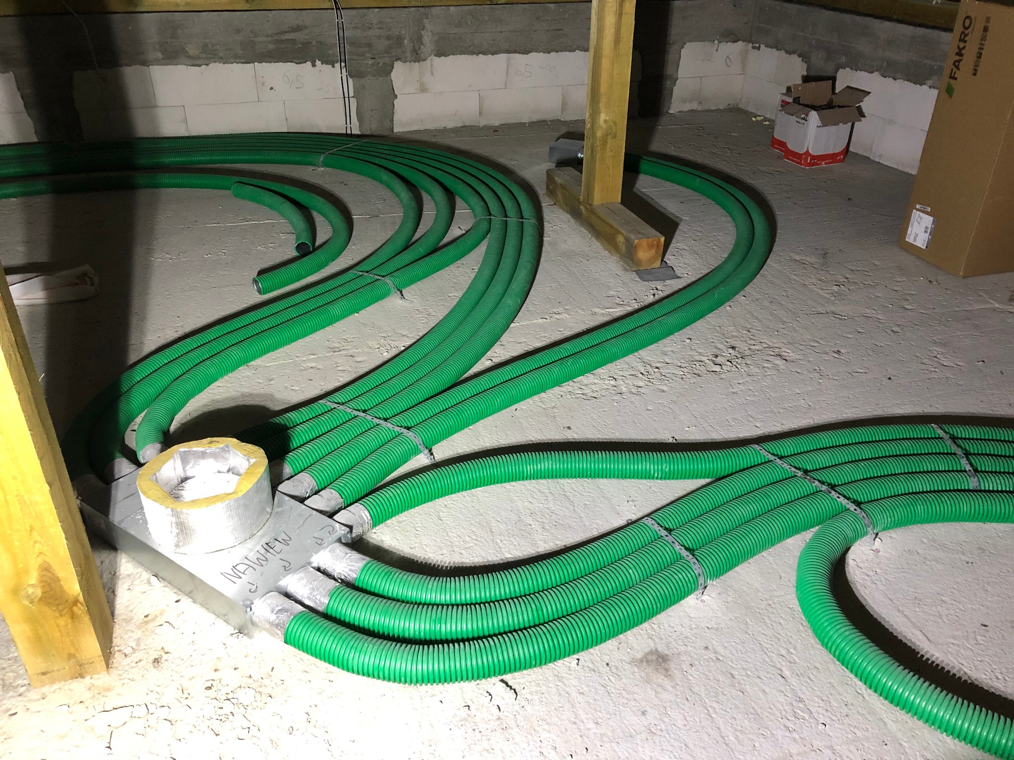 Zielone, karbowane rury wentylacyjne rozprowadzone na podłodze w surowym wnętrzu, połączone metalową płytą z izolacją, widoczne drewniane elementy konstrukcyjne i kartony.