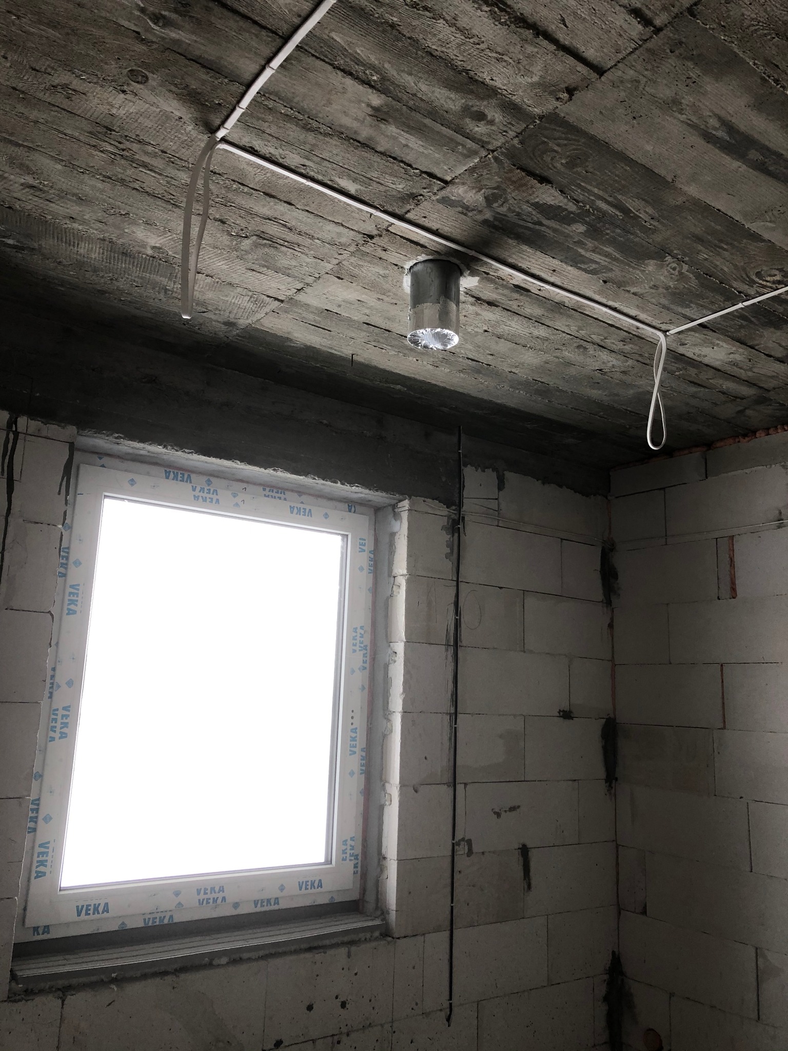 Surowy stan wnętrza z widocznym oknem VEKA, betonowym stropem z instalacją elektryczną i puszką montażową, ściany z betonu komórkowego przed wykończeniem.