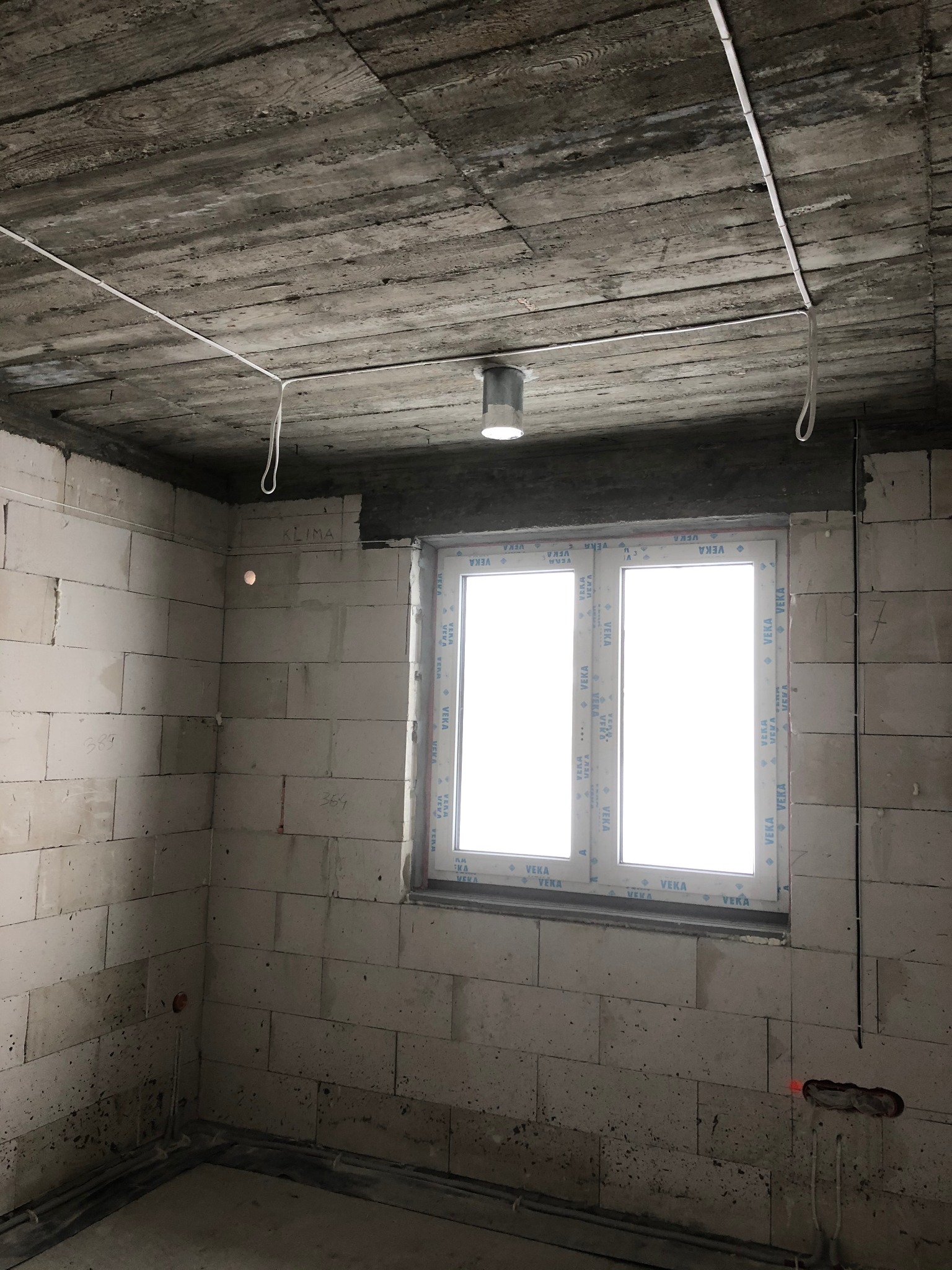 Surowy stan wnętrza w trakcie budowy lub remontu. Widoczne ściany z bloczków, okno PCV z naklejkami producenta, sufit betonowy z widocznymi przewodami elektrycznymi i pojedynczym punktem...