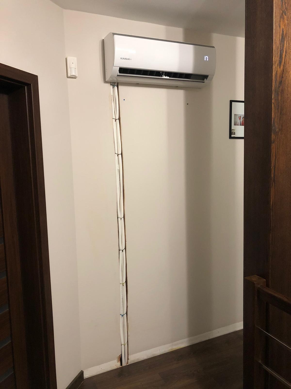 Klimatyzator Kaisai zamontowany na ścianie w pokoju, widoczne białe kable w prowadnicy ściennej, brązowe drewniane elementy wystroju.