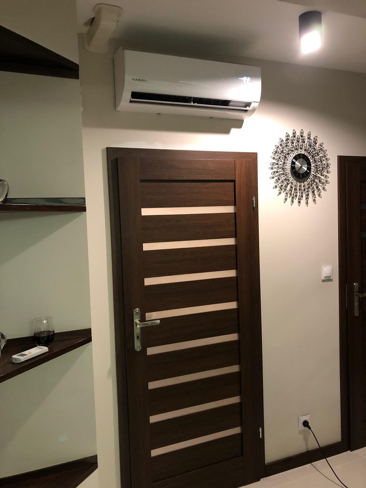 Biały klimatyzator Kaisai zamontowany nad brązowymi drzwiami z poziomymi paskami w nowoczesnym wnętrzu, obok dekoracyjny zegar słoneczny na ścianie.