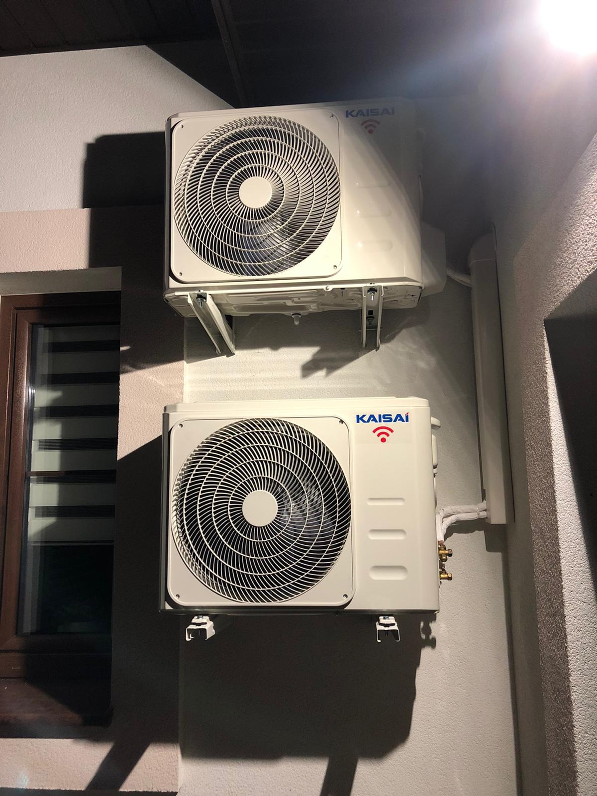 Dwa klimatyzatory Kaisai zamontowane jeden nad drugim na zewnętrznej ścianie budynku, widoczne okno z żaluzjami w tle.