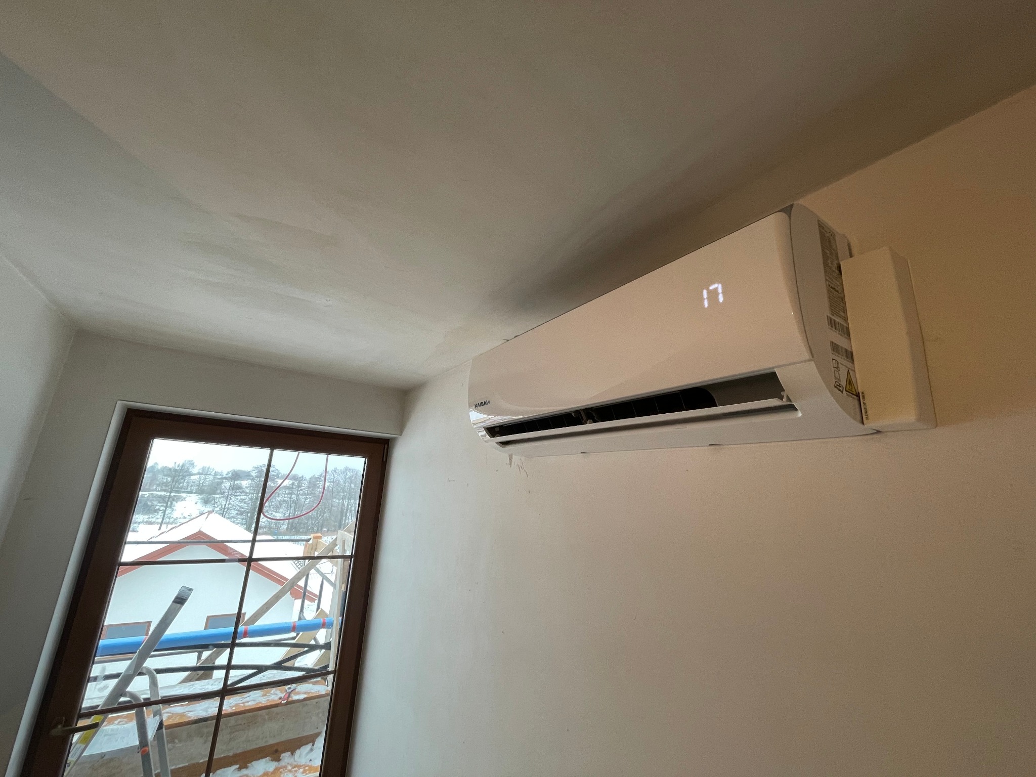 Biały klimatyzator ścienny marki Kaisai z wyświetlaną temperaturą 17 stopni, zamontowany w pomieszczeniu w trakcie remontu, widok na zewnątrz przez okno z rusztowaniem i śniegiem.