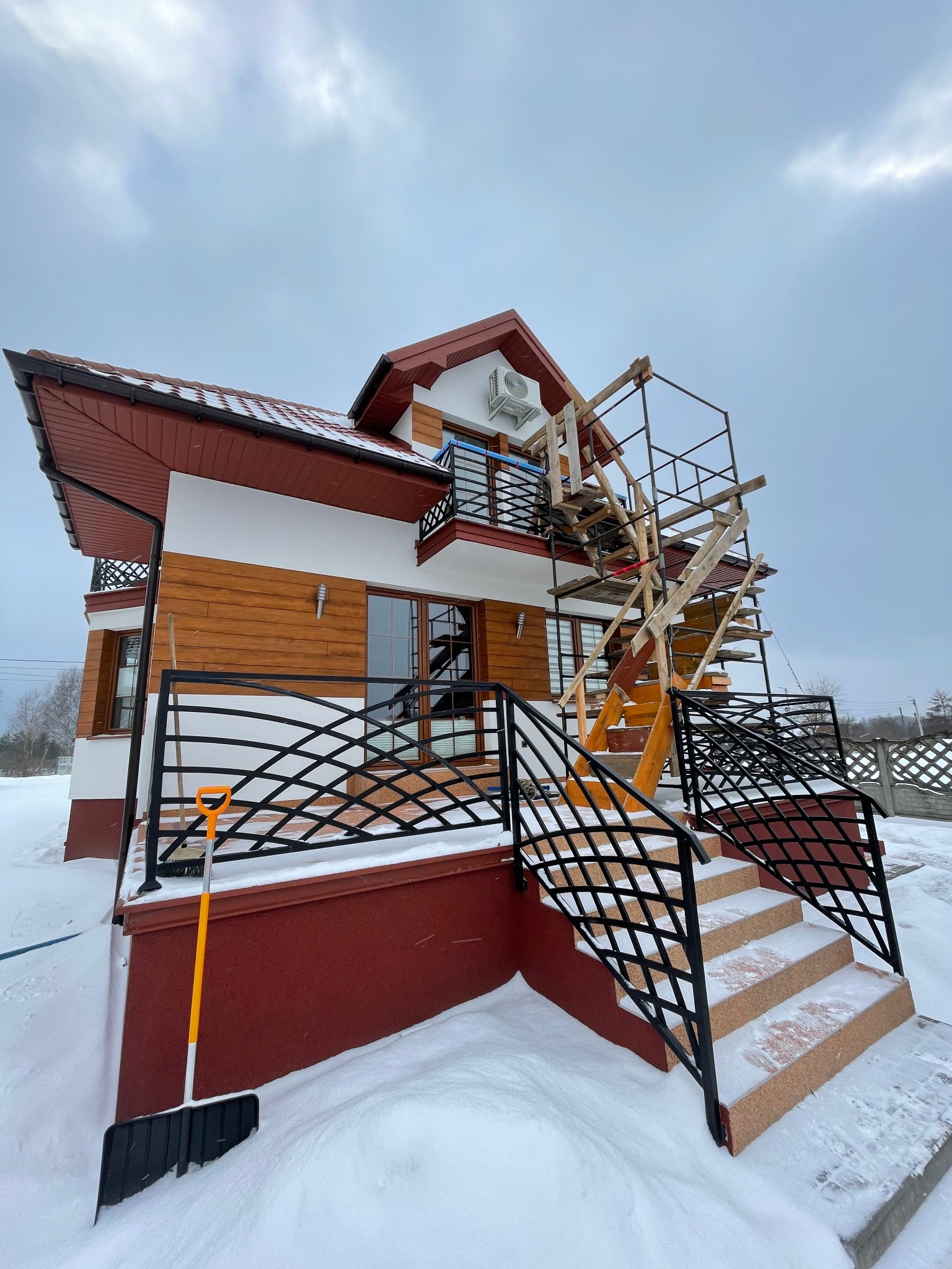 Dwupiętrowy dom z drewnianą elewacją i balkonem, na którym zamontowano klimatyzator. Widoczne rusztowanie i odśnieżona łopata przed schodami wejściowymi pokrytymi śniegiem.