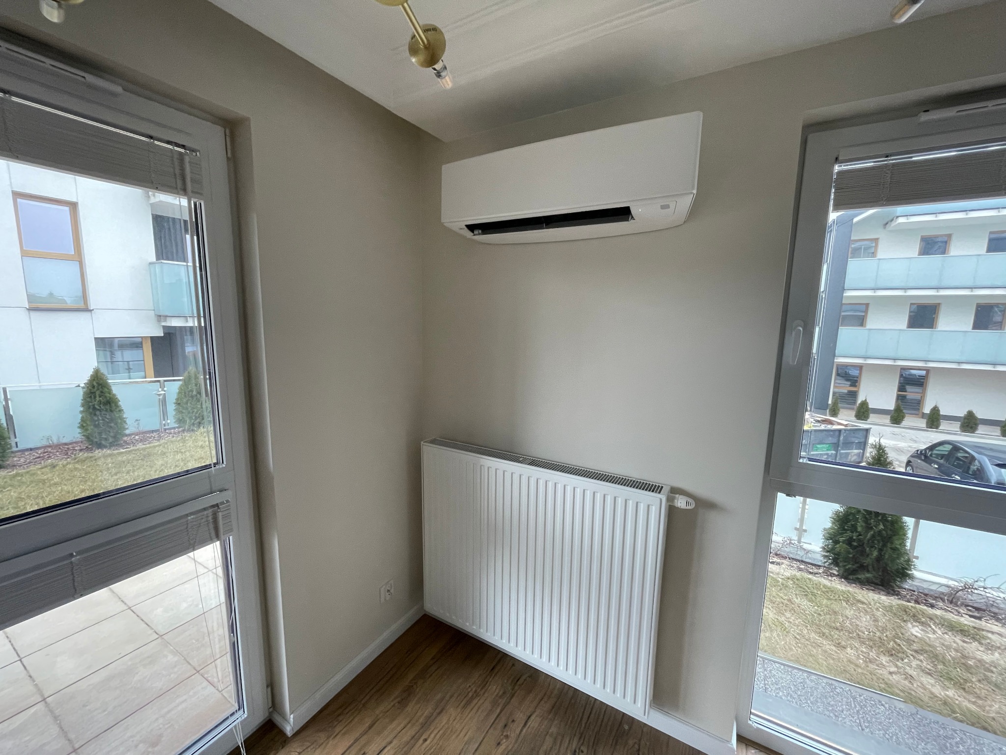 Biały klimatyzator ścienny zamontowany w rogu pokoju, widoczny grzejnik pod spodem, dwa okna z widokiem na zewnątrz, drewniana podłoga, minimalistyczny design.