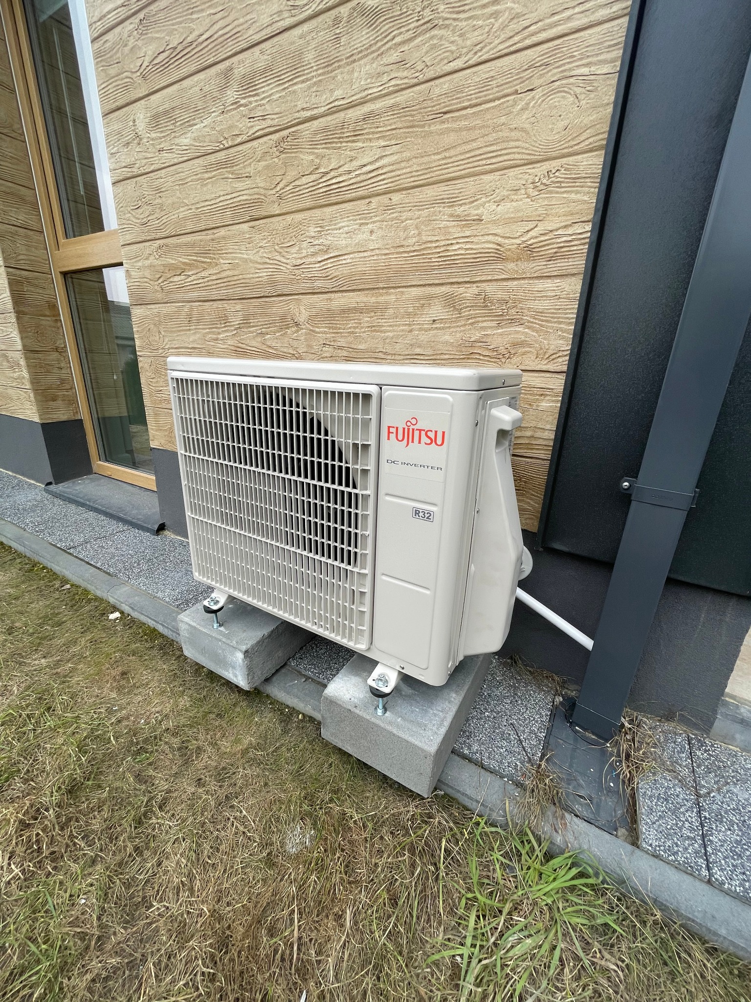 Zewnętrzna jednostka klimatyzacji Fujitsu zamontowana na betonowych blokach przy ścianie budynku z drewnianą elewacją.