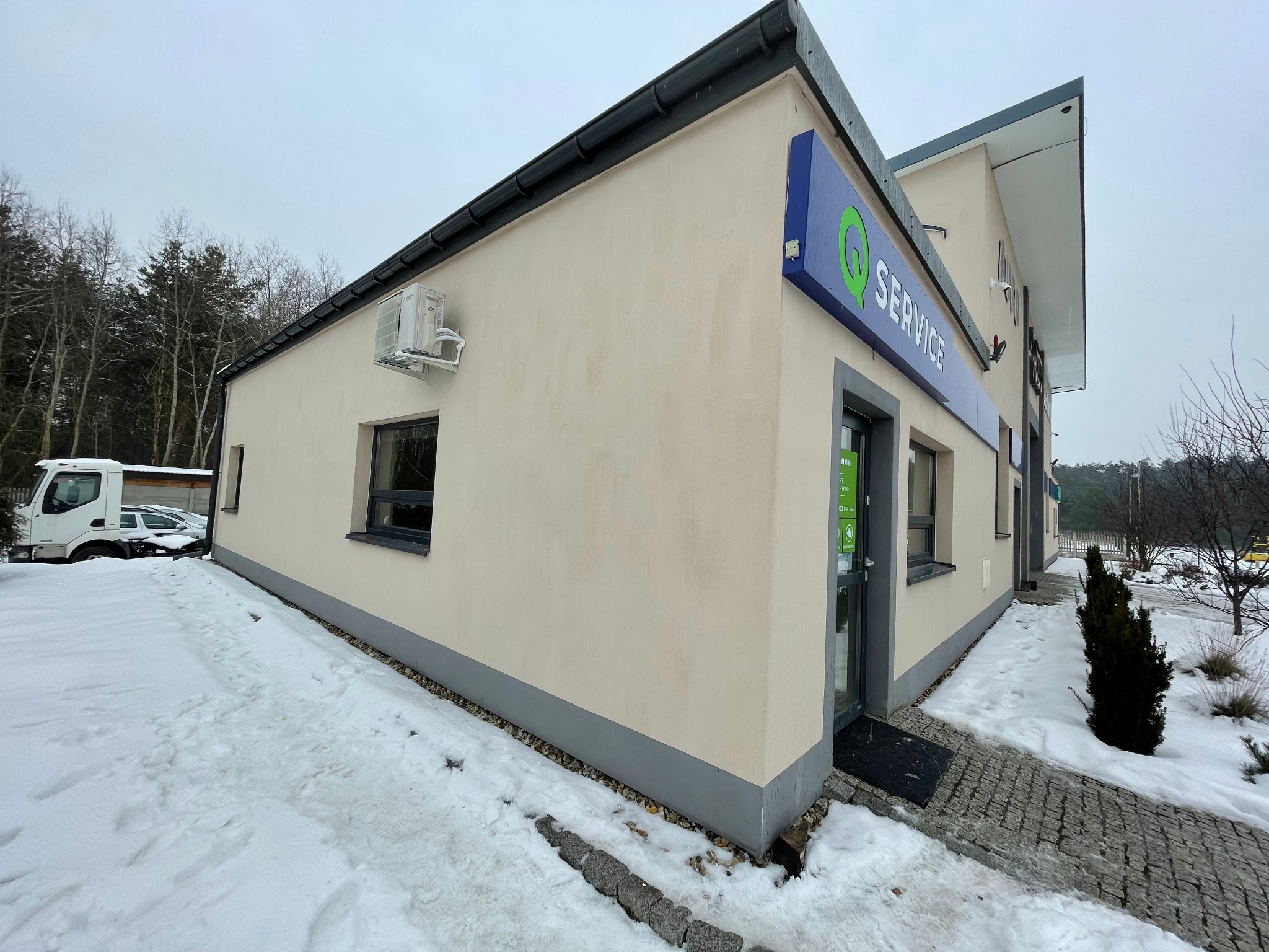 Zewnętrzny widok budynku usługowego z zamontowaną klimatyzacją na ścianie, w zimowej scenerii ze śniegiem na ziemi i firmowym szyldem nad wejściem.