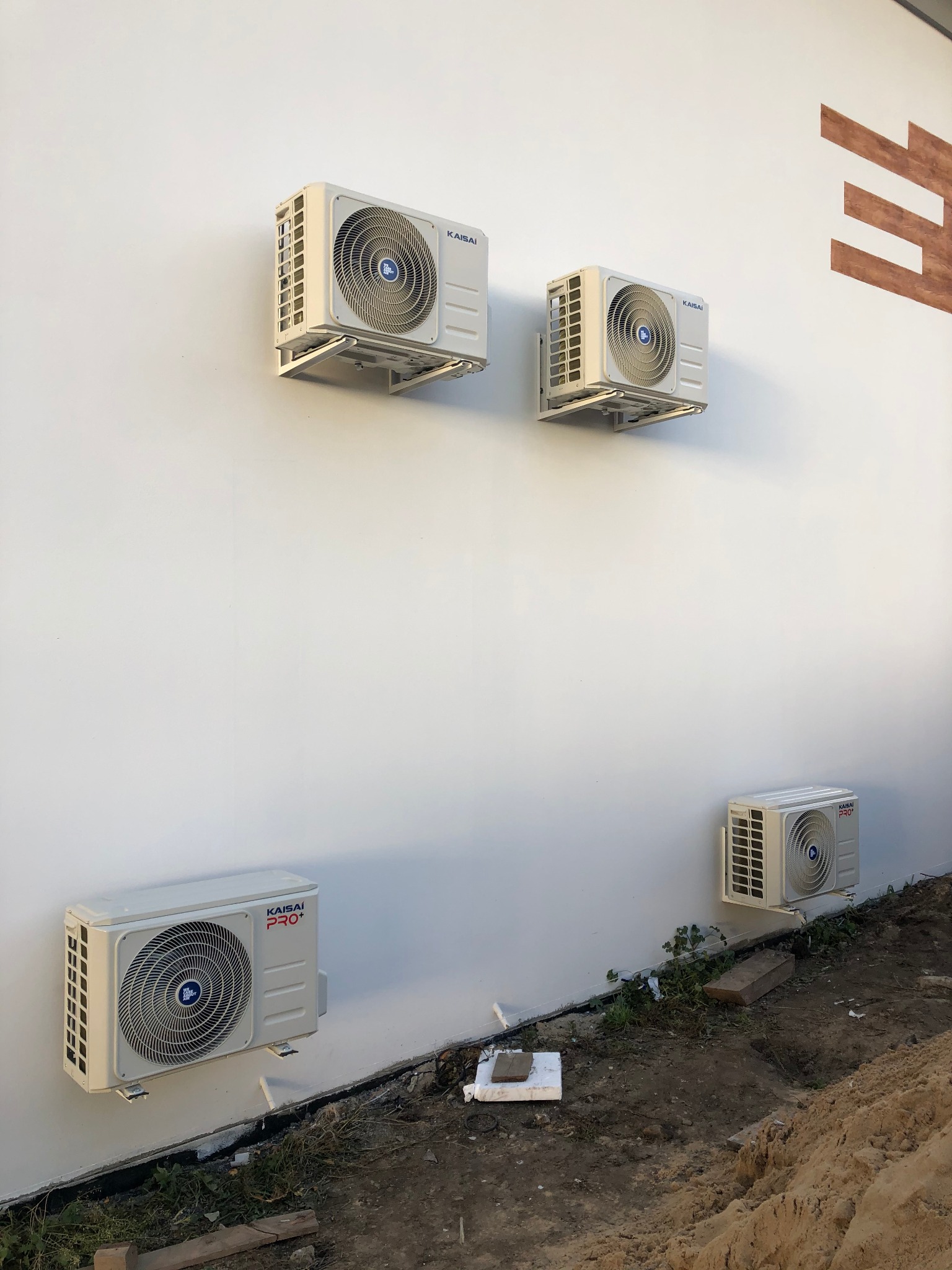 Cztery klimatyzatory Kaisai zamontowane na białej ścianie budynku, dwa górne z widocznymi wspornikami, dolny rząd blisko ziemi z widocznym nierównym terenem i fragmentami roślinności.