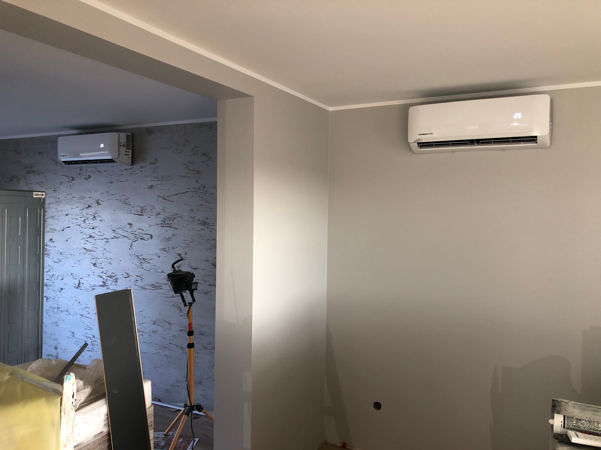 Biała jednostka klimatyzacji Kaisai Pro zamontowana na szarej ścianie w jasnym pomieszczeniu, widoczna druga jednostka w przejściu, lampa na statywie i materiały budowlane w tle.