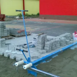 Nierównomiernie ułożona szara kostka brukowa na piasku, z niebieskim urządzeniem do wyrównywania podłoża, paleta z czerwoną kostką w tle, widok z góry.