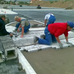 Czterech mężczyzn w roboczych ubraniach układa drobną kostkę granitową na przygotowanym podłożu, widoczne elementy betonowe i narzędzia, w tle plac budowy.