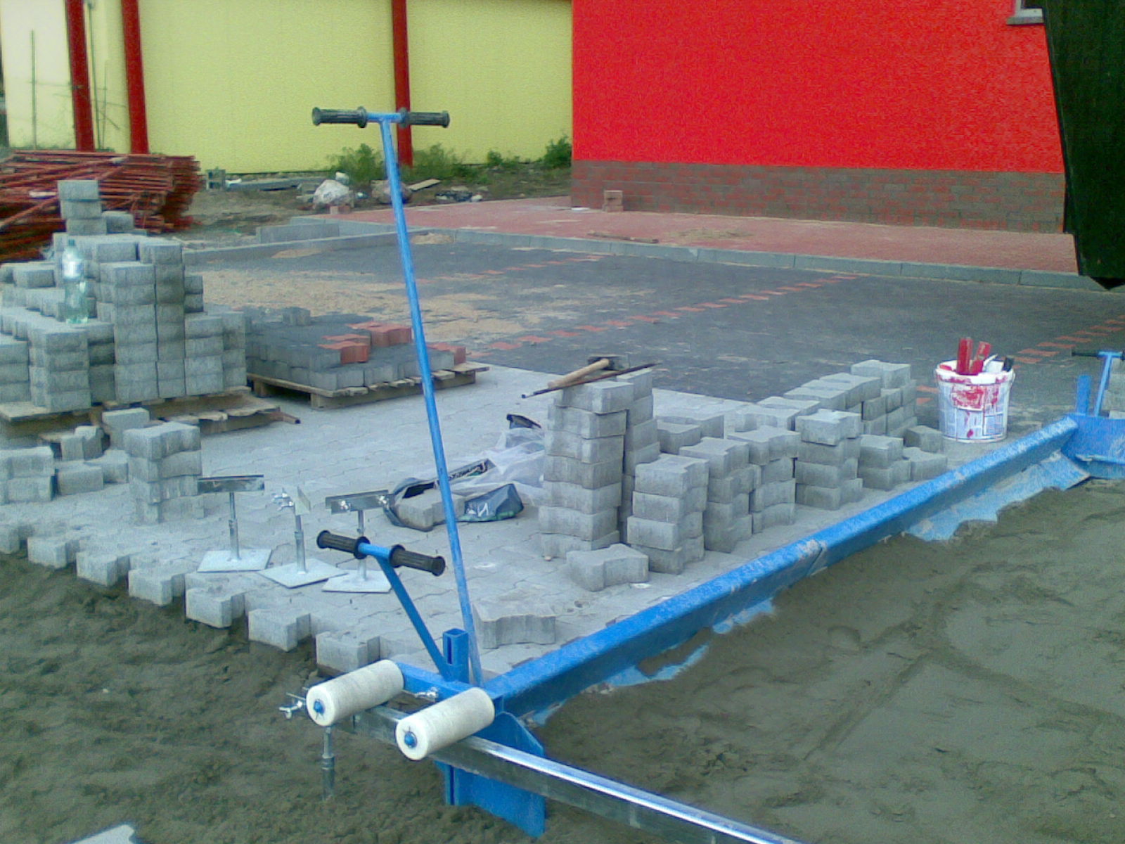 Nierównomiernie ułożona szara kostka brukowa na piasku, z niebieskim urządzeniem do wyrównywania podłoża, paleta z czerwoną kostką w tle, widok z góry.