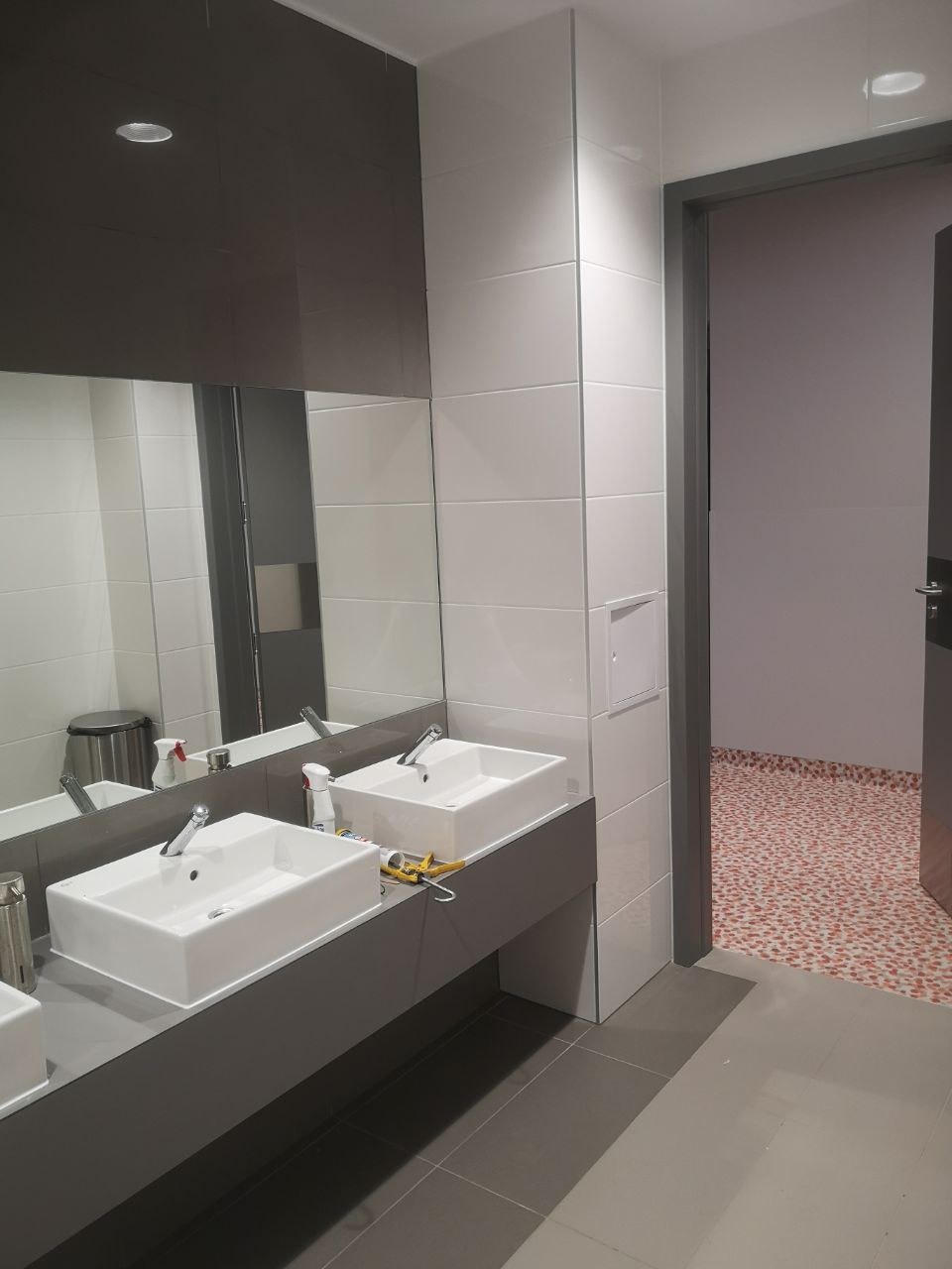 Nowoczesna łazienka publiczna z dwoma białymi umywalkami prostokątnymi na szarym blacie, białe płytki na ścianach, szare płytki podłogowe i mozaika na podłodze w dalszej części pomieszczenia.