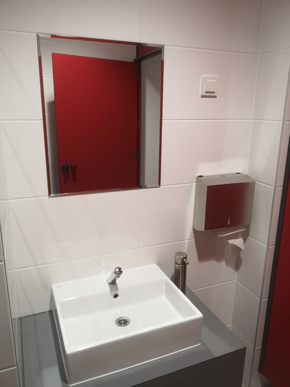 Nowoczesna łazienka publiczna: biała, kwadratowa umywalka nablatowa z chromowaną baterią, dozownik mydła w kolorze stali nierdzewnej, podajnik ręczników papierowych, lustro z odbiciem czerwonych...