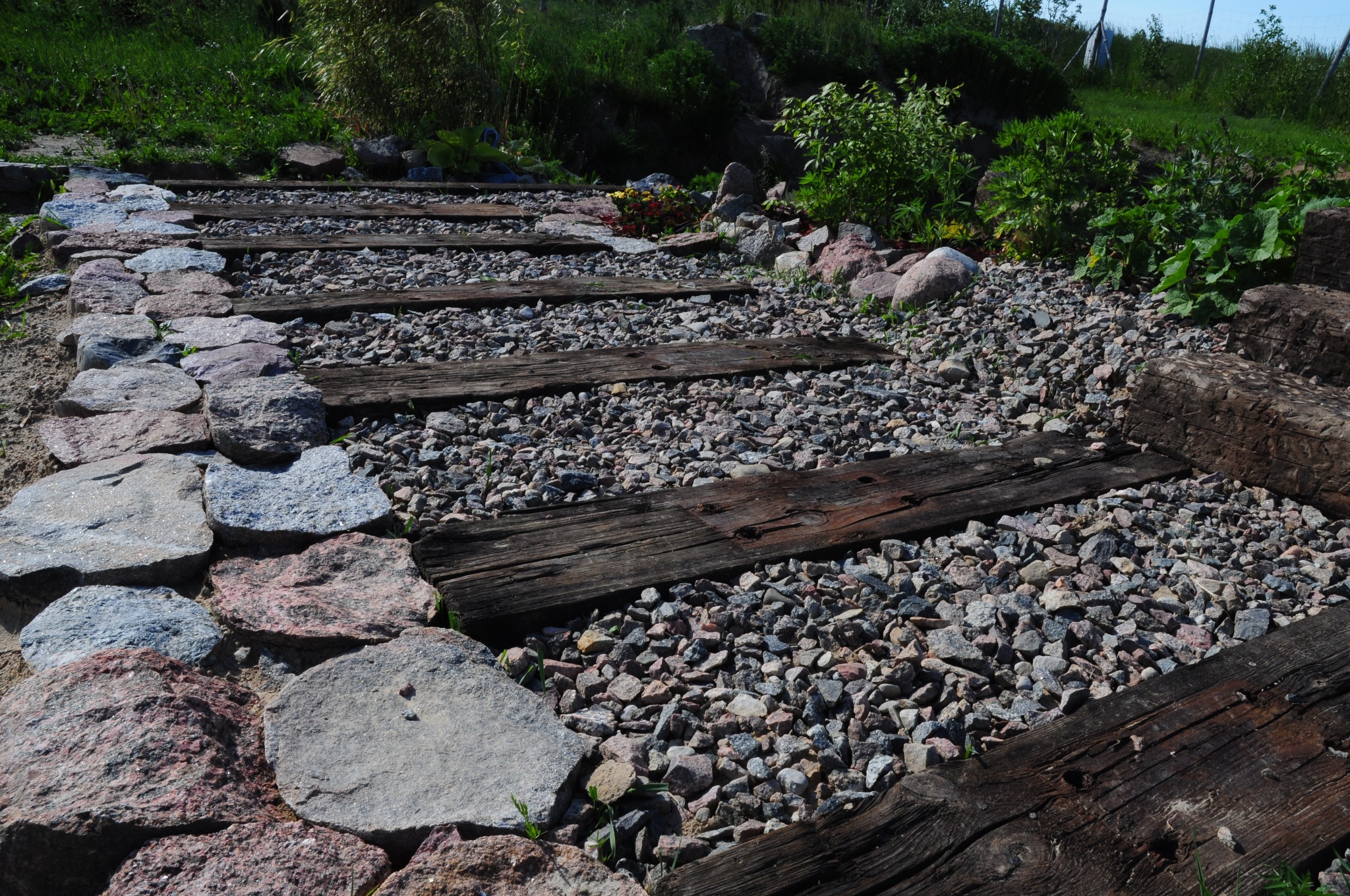 Ścieżka w ogrodzie wykonana z nieregularnych kamieni i drewnianych bali, wysypana drobnym żwirem, z zielenią w tle.