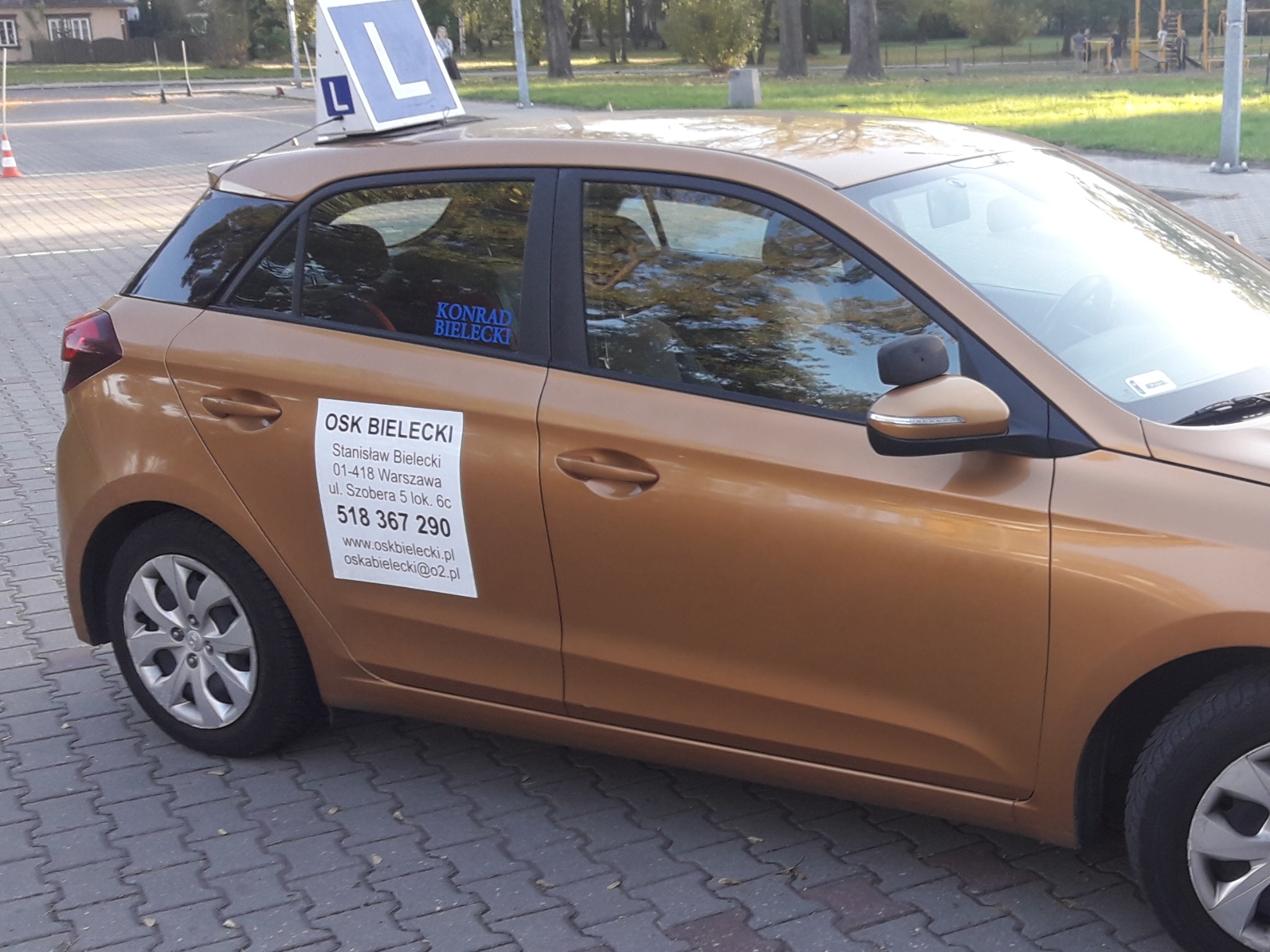 Złoty samochód z naklejką OSK Bielecki i znakiem 'L' na dachu, zaparkowany na chodniku z kostki brukowej, widok z boku.