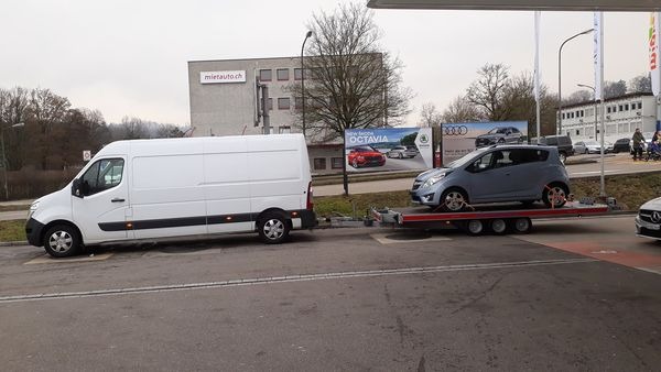 Biały samochód dostawczy Renault Master z przyczepą lawetą, na której przewożony jest szary Opel Karl, na tle budynku z logo Mietauto.ch i billboardu z samochodami Skoda Octavia.