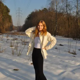 Portret młodej kobiety w białej kurtce i czarnych spodniach na tle ośnieżonego lasu z widoczną linią wysokiego napięcia w oddali.