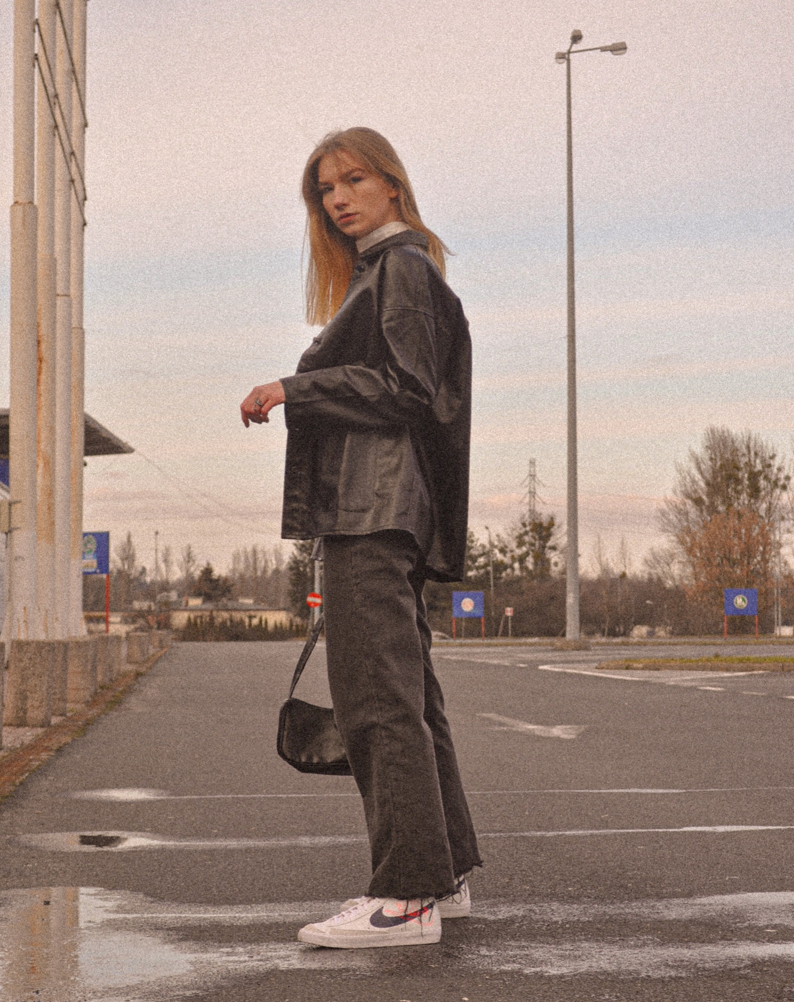 Kobieta w czarnej, skórzanej kurtce i szarych dżinsach pozuje na mokrym parkingu, trzymając czarną torebkę na ramieniu.