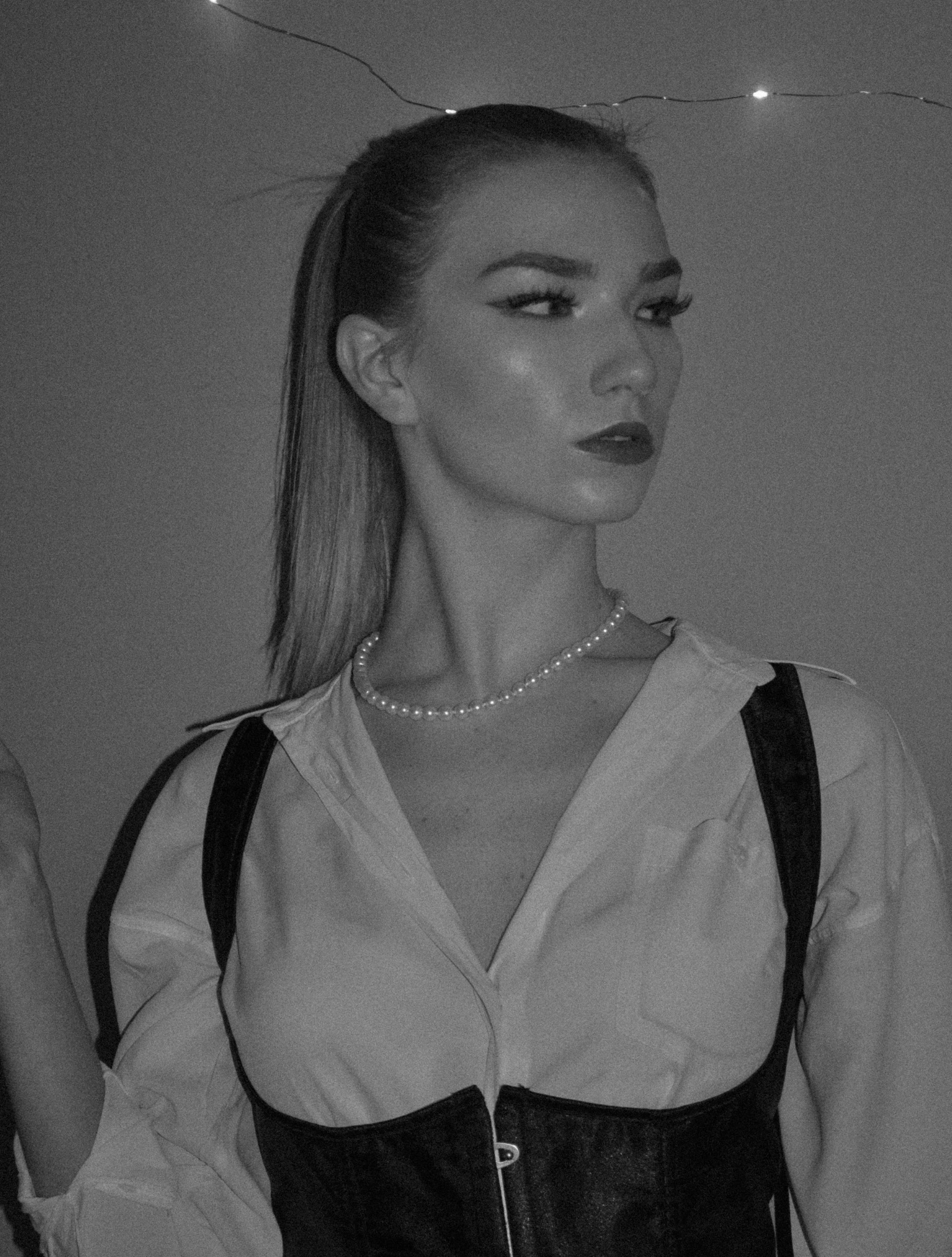 Czarno-białe portretowe ujęcie kobiety z makijażem, w koszuli z rozpiętym kołnierzykiem, perłowym naszyjnikiem i czarnym gorsecie na szelkach, oświetlonej lampkami w tle.