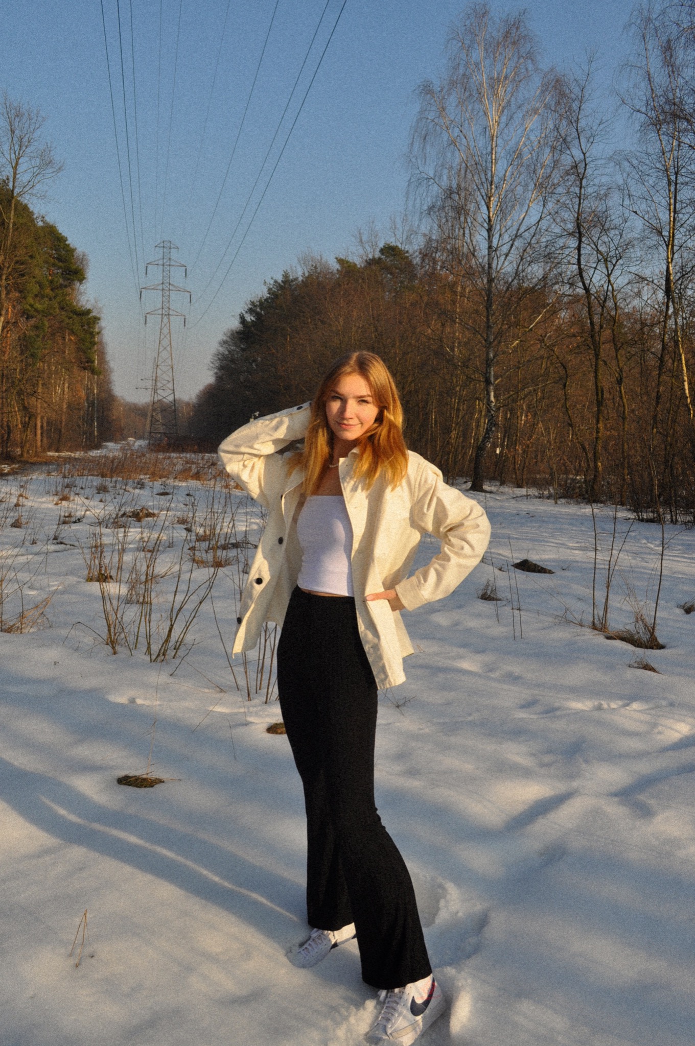 Portret młodej kobiety w białej kurtce i czarnych spodniach na tle ośnieżonego lasu z widoczną linią wysokiego napięcia w oddali.