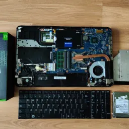 Otwarty laptop z widoczną płytą główną, dyskiem SSD Goodram CL100 120GB, wyjętym dyskiem twardym Toshiba, napędem DVD, klawiaturą oraz pudełkiem po baterii Green Cell Fresh Energy.