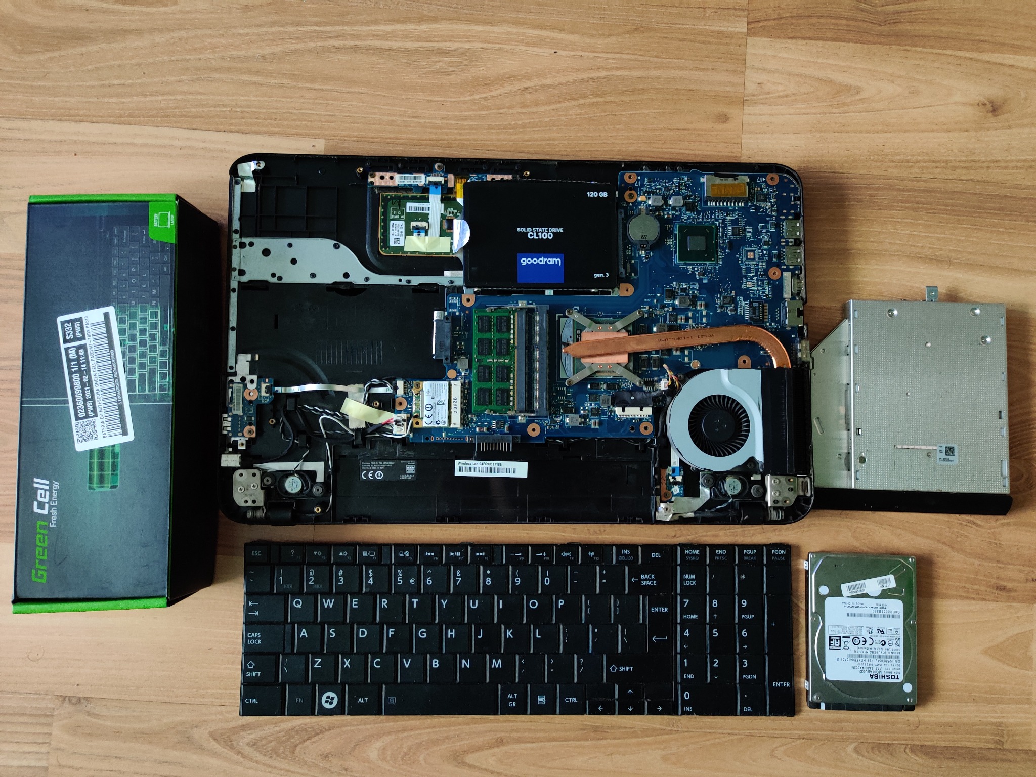 Otwarty laptop z widoczną płytą główną, dyskiem SSD Goodram CL100 120GB, wyjętym dyskiem twardym Toshiba, napędem DVD, klawiaturą oraz pudełkiem po baterii Green Cell Fresh Energy.