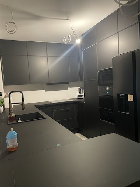 Nowoczesna czarna kuchnia z matowymi frontami, widoczne niedokończone podłączenie oświetlenia sufitowego i butelka dla dziecka na blacie.