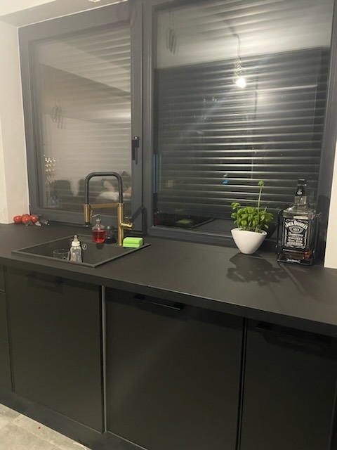 Nowoczesna, czarna zabudowa kuchenna pod oknem, z czarnym zlewem i złotą baterią, uzupełniona o butelkę Jack Daniel's i doniczkę z bazylią.