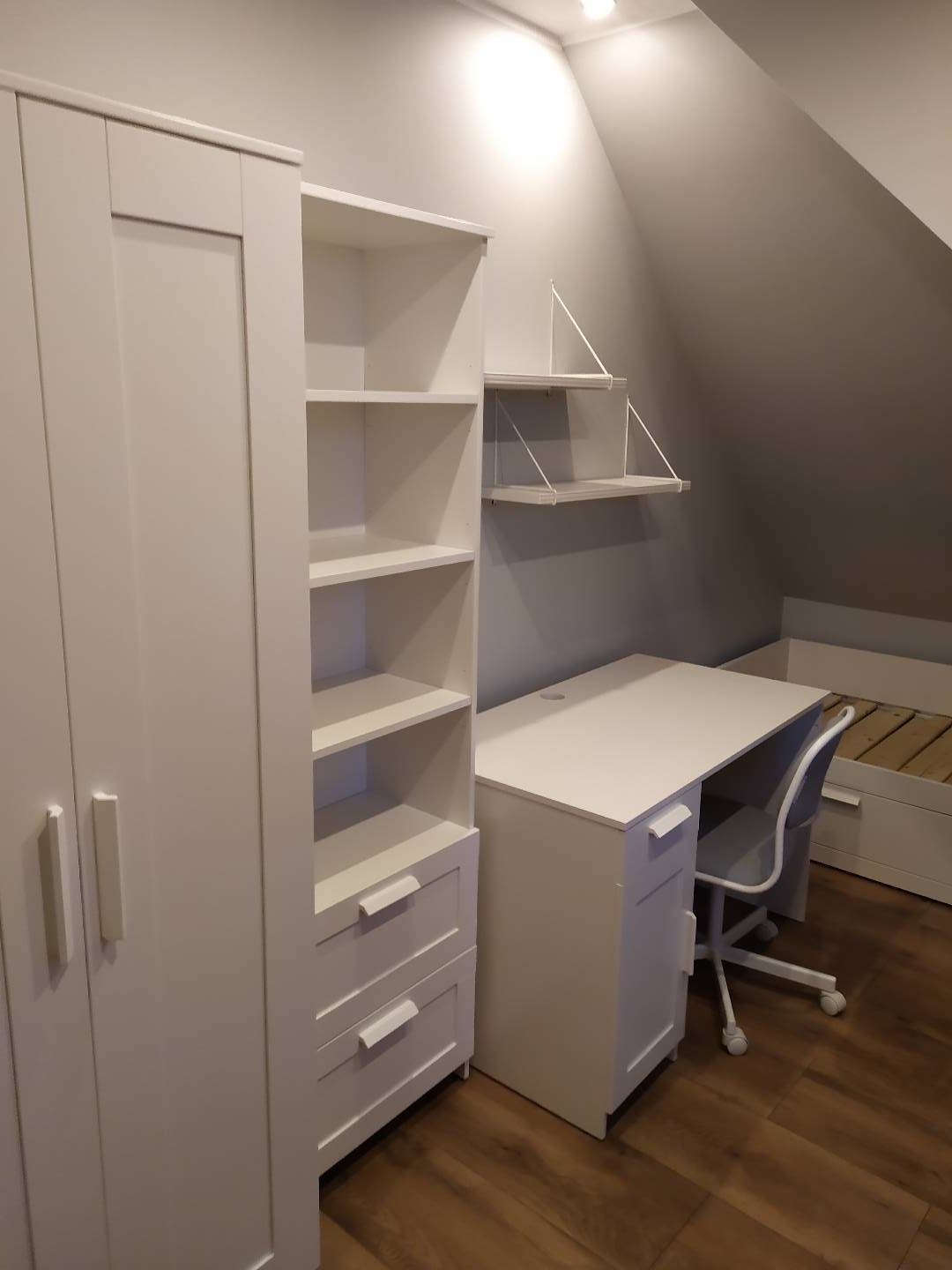 Komplet białych mebli w pokoju na poddaszu: szafa, regał z szufladami, biurko z krzesłem i półki wiszące.