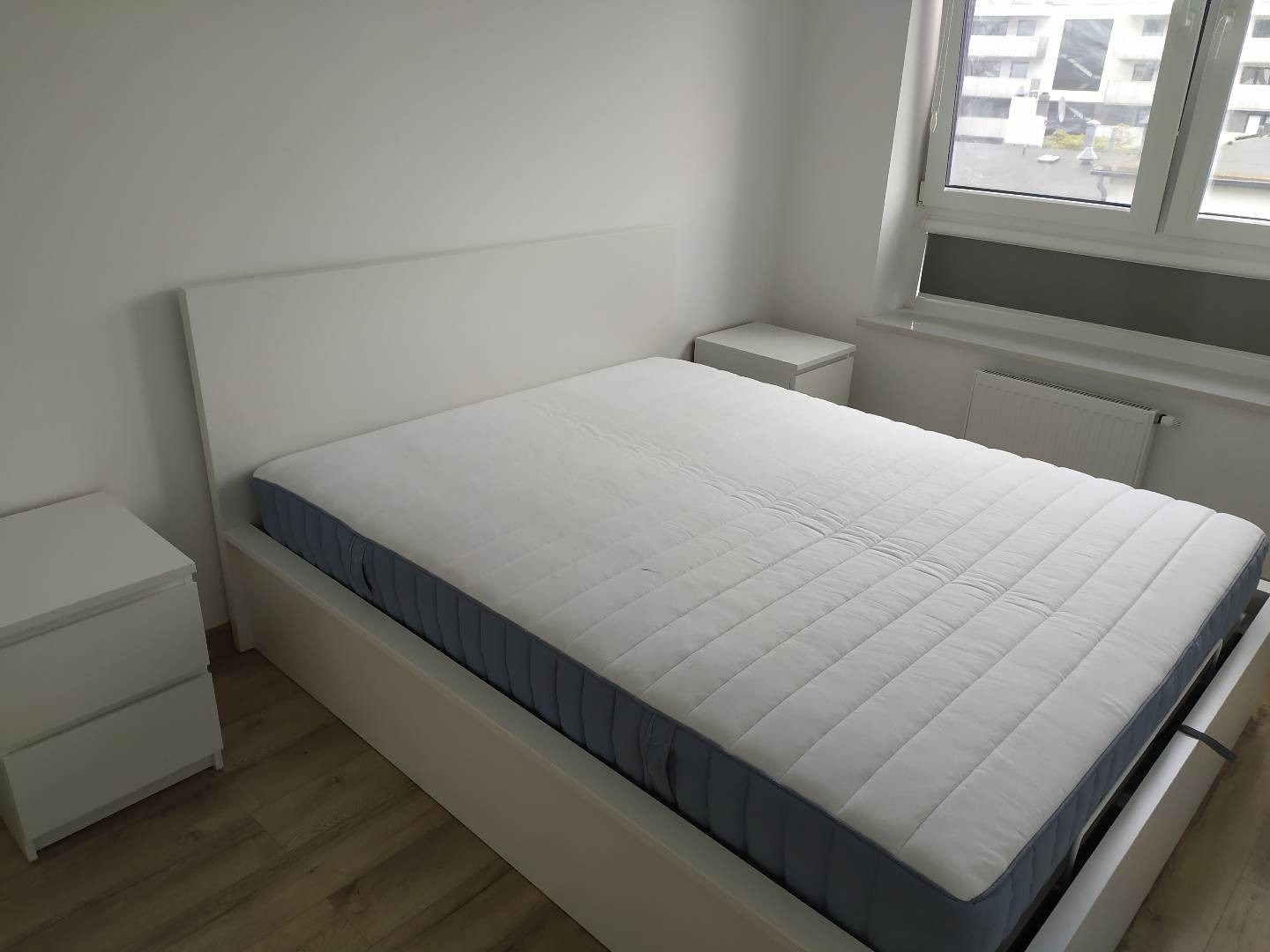 Nowoczesna, biała rama łóżka z materacem w minimalistycznej sypialni, widoczne dwie białe szafki nocne i okno z widokiem na budynki.