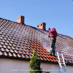 Pracownik w czerwonej bluzie i zielonych spodniach, zabezpieczony uprzężą, czyści dach z dachówki ceramicznej za pomocą myjki ciśnieniowej, stojąc na aluminiowej drabinie. Część dachu jest już...