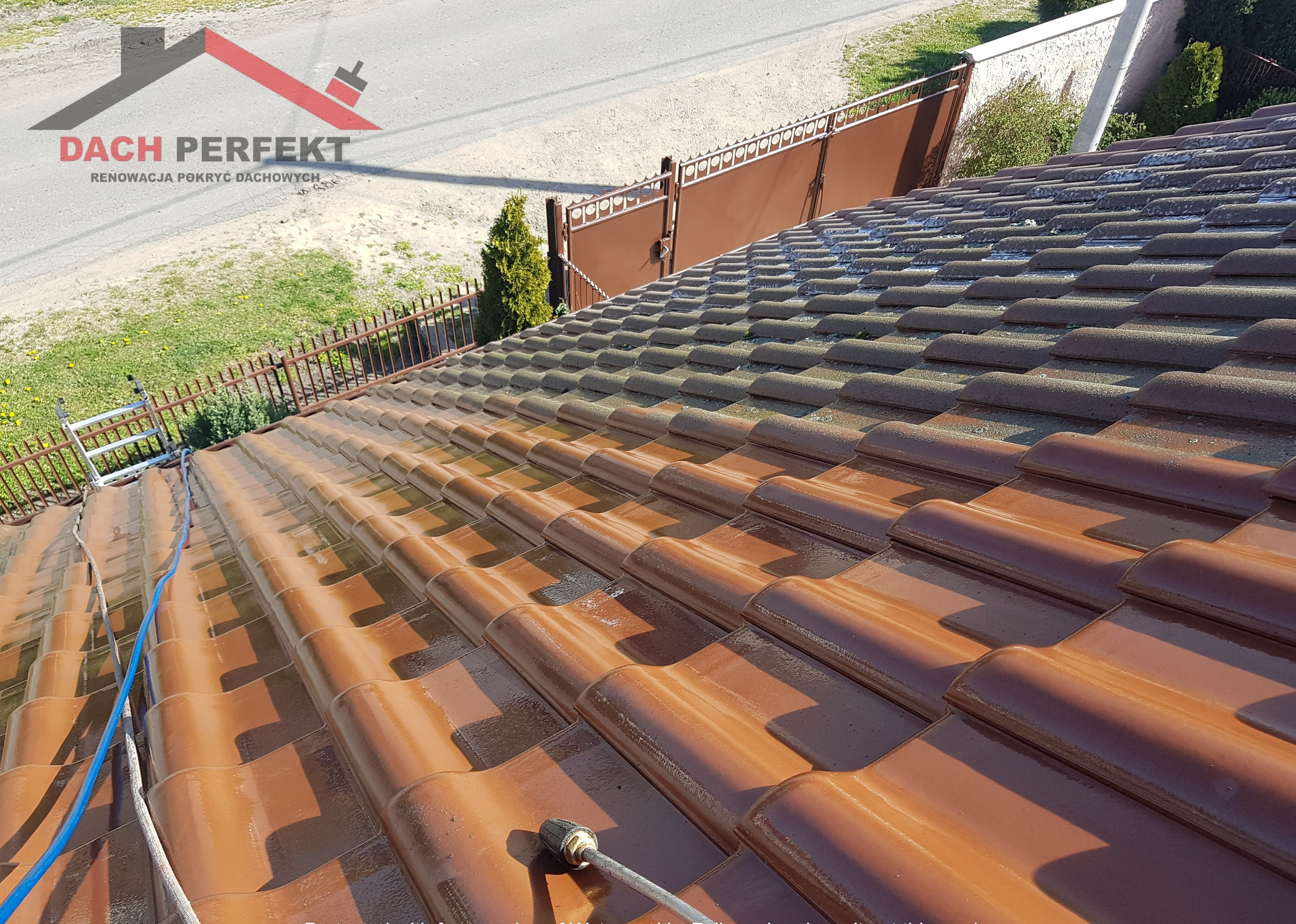 Częściowo umyty dach z brązowej dachówki ceramicznej, widoczna różnica w kolorze między czystą a brudną powierzchnią, wąż ciśnieniowy i lanca do mycia w dolnej części kadru, w tle brama posesji...