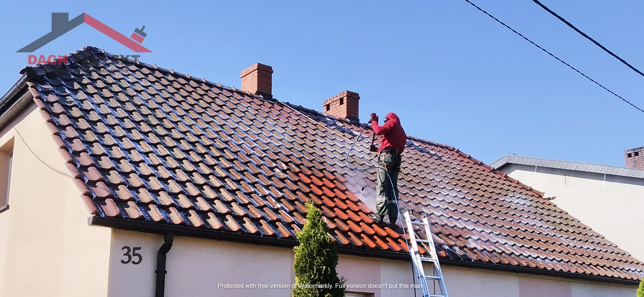 Pracownik w czerwonej bluzie i zielonych spodniach, zabezpieczony uprzężą, czyści dach z dachówki ceramicznej za pomocą myjki ciśnieniowej, stojąc na aluminiowej drabinie. Część dachu jest już...