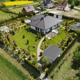 Widok z lotu ptaka na zadbany ogród z nowoczesnym domem, ścieżką z kostki, pergolą, licznymi nasadzeniami ozdobnymi i równo przystrzyżonym trawnikiem, otoczony tujami i metalowym ogrodzeniem.