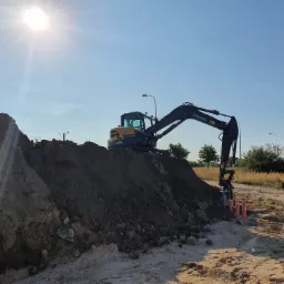 Żółto-czarna koparka Volvo na placu budowy, ustawiona na wzniesieniu ziemi, z trzema pomarańczowymi rurami wystającymi z ziemi, oświetlona jasnym słońcem.