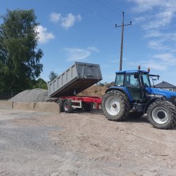 Uslugi ogólnobudowlane Jakub Kowalik - Niebieski traktor z przyczepą wywrotką unosi skrzynię ładunkową, wysypując żwir na placu budowy w słoneczny dzień.