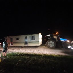 Uslugi ogólnobudowlane Jakub Kowalik - Ciężki traktor New Holland holuje mobilny domek letniskowy w nocy; widoczna sylwetka mężczyzny i światła samochodu w tle.